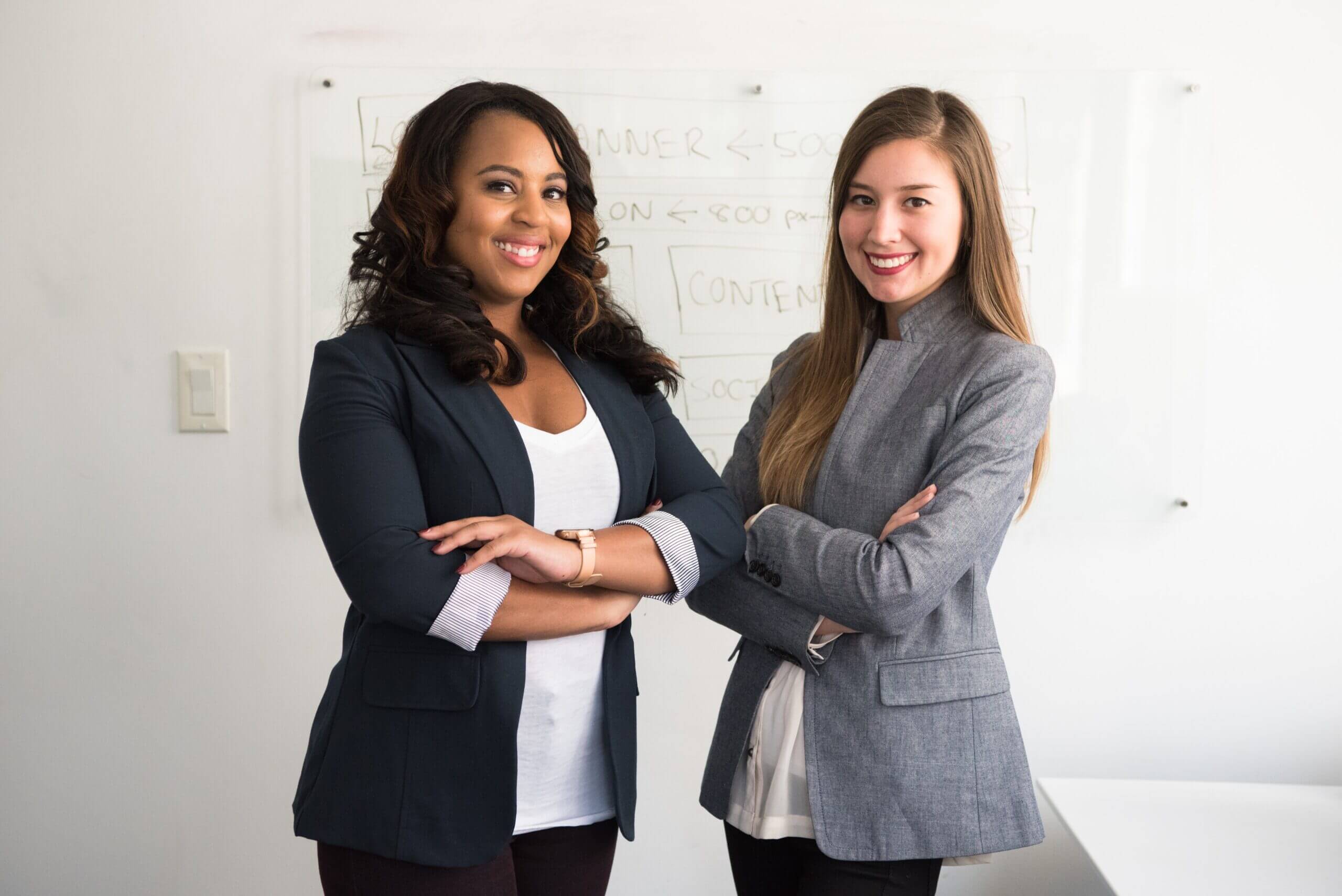 Two professional women smiling confidently in front of a whiteboard with planning notes.