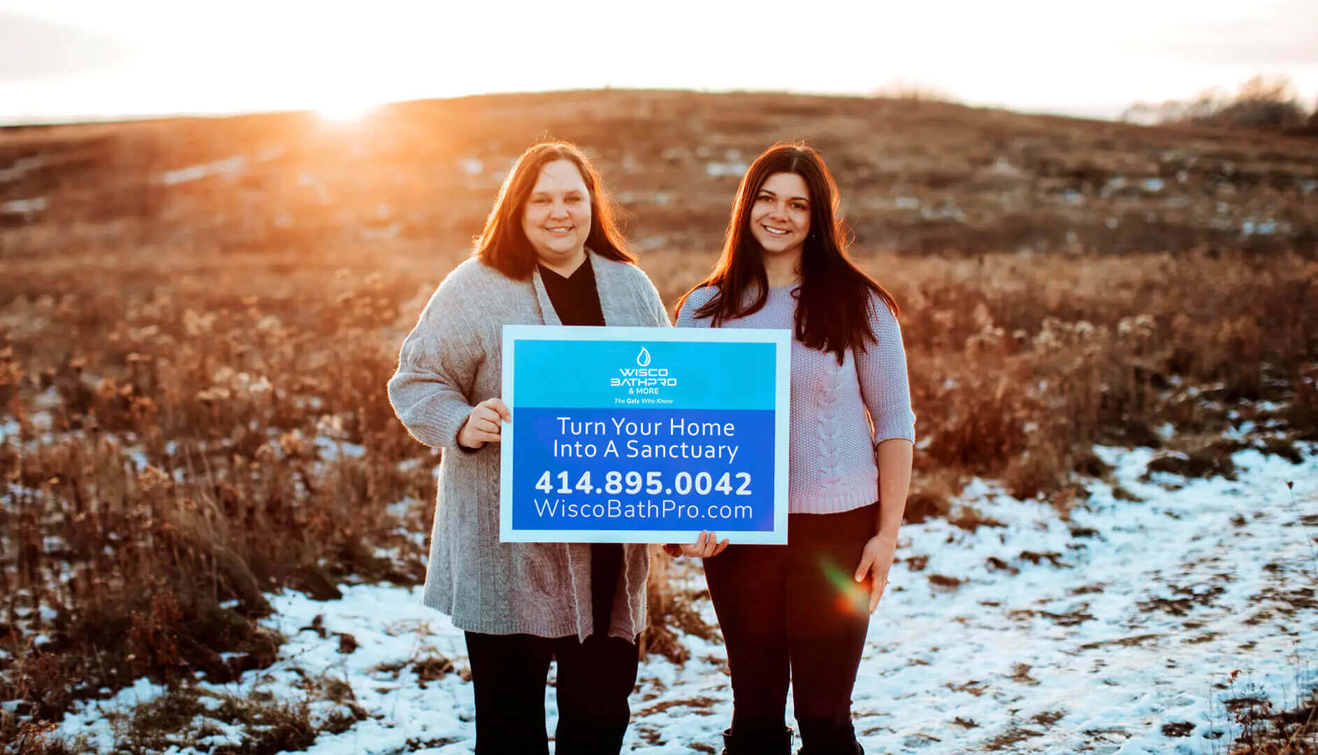 <br />
The image shows two women standing in a field at sunset, holding a blue sign with the logo and contact information for "Wisco BathPro & More". The sign reads "Turn Your Home Into A Sanctuary" along with a phone number and website. The field has sparse vegetation and patches of snow, and the warm glow of the sun highlights the friendly and inviting expressions of the women.