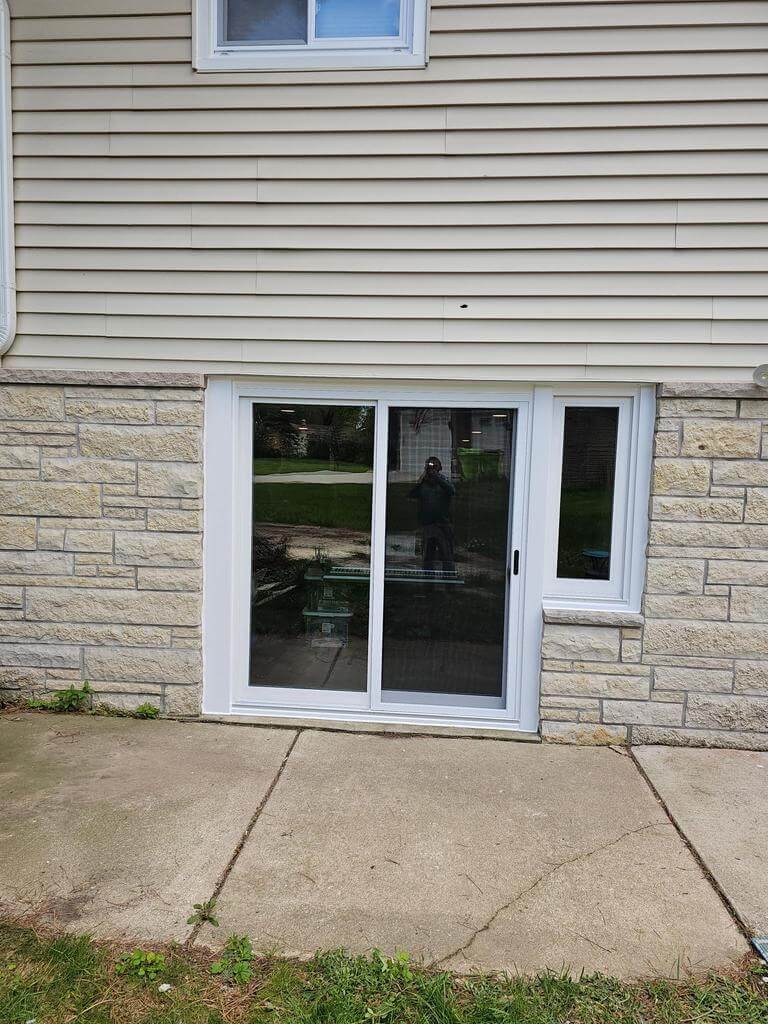 basement-sliding-door-installation Newly installed white sliding glass door on a basement-level exterior with stone and vinyl siding.