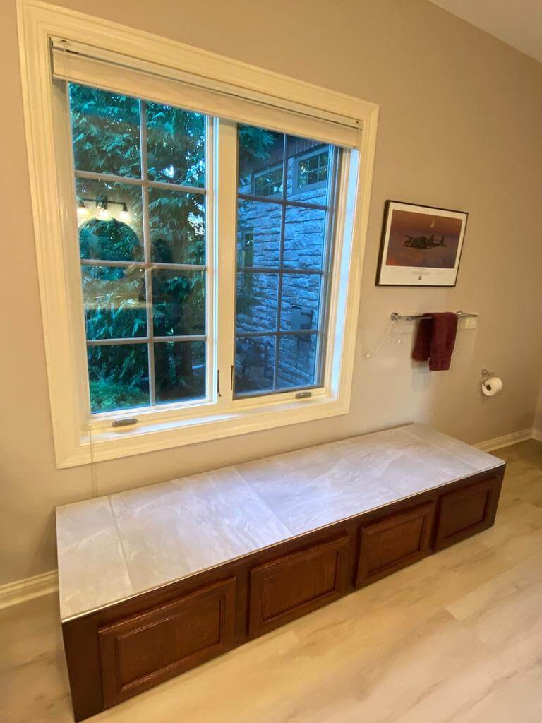bathroom-window-bench-storage Large bathroom window with a built-in bench featuring marble top and wood storage cabinets underneath.