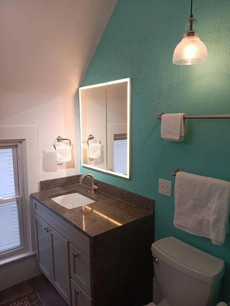 teal-bathroom-vanity-lighted-mirror Modern bathroom with a teal accent wall, granite countertop vanity, lighted mirror, and pendant light above the toilet.